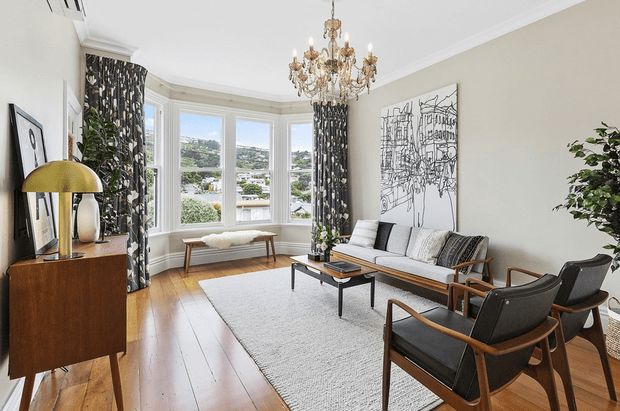 A bright, refined living room with a bay window overlooking a residential landscape. The room features polished timber flooring, a light neutral palette, patterned floor-to-ceiling curtains, a vintage-style chandelier, mid-century-inspired timber and black leather lounge chairs, a pale sofa, a low black coffee table, a textured rug, and a dark timber sideboard.
