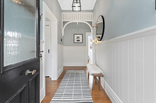 A bright, refined residential hallway viewed from an open dark-painted front door. The corridor features warm timber flooring, white vertical wall panelling with a chair rail, pale blue-grey upper walls, white trim, an arched opening with decorative moulding, a striped runner, a timber bench, an oval wall mirror, framed artwork, and a hanging lantern-style light.