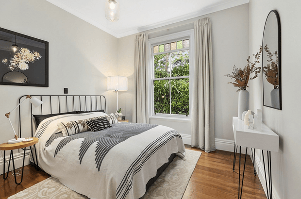A refined bedroom with a black metal bed frame, layered cream and grey bedding, pale neutral walls, timber flooring, full-length curtains, and a large sash-style window overlooking greenery. A slim white console, arched wall mirror, bedside table, floor lamp, framed artwork, and patterned rug complete the calm, contemporary interior.