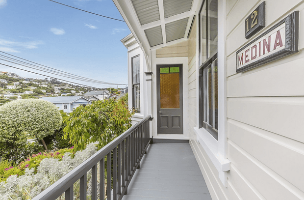A bright exterior veranda or balcony walkway along a traditional weatherboard building. The covered porch has white horizontal cladding, grey-painted timber flooring and balustrades, tall timber-framed windows, and a grey door with a textured amber glass panel. Vintage-style signs reading “12” and “MEDINA” are mounted on the wall. A landscaped garden and hillside neighbourhood are visible beyond the railing.
