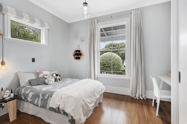 Bright, compact bedroom with soft grey walls, white trim, timber flooring, and a large curtained window. The room features a double bed with layered grey and white bedding, a small bedside table, playful wall decor, and a simple white desk and chair.