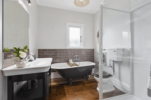 A bright, compact traditional bathroom with a freestanding black-and-white clawfoot bathtub, white pedestal-style basin vanity, enclosed glass shower, toilet, and a large wall mirror. The room features white painted walls, a dark brown brick-pattern tiled dado, polished timber flooring, chrome fixtures, and a small frosted window.