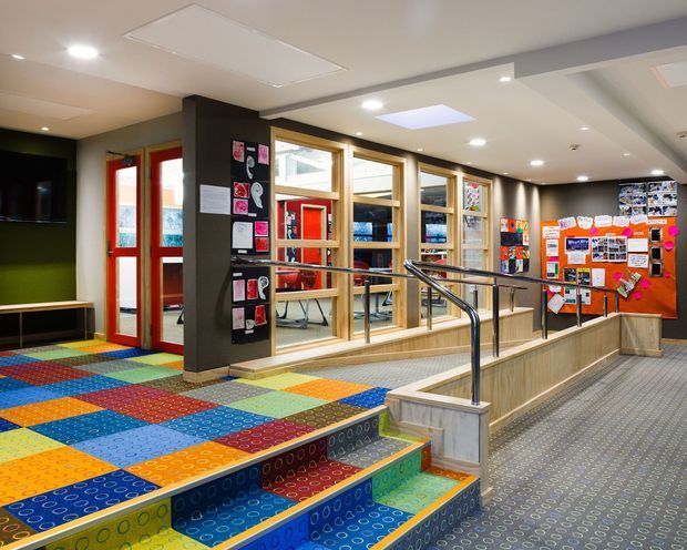 Interior view of a colourful educational or community facility, featuring a patterned multicolour carpeted floor and steps, a ramp with stainless-steel handrails, timber-framed interior glazing, red doors, display boards, and recessed ceiling lighting.