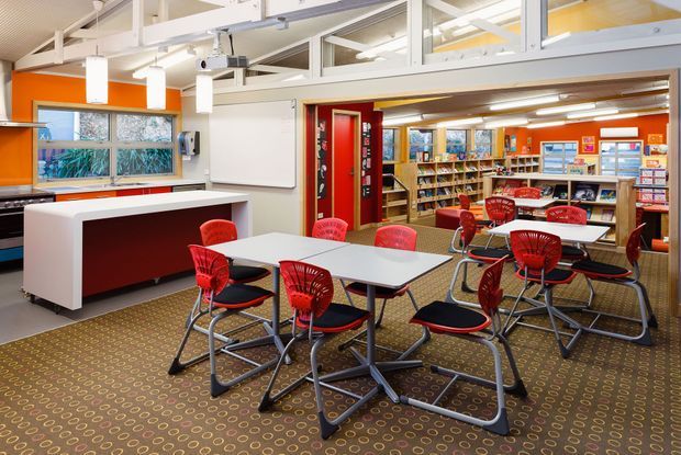 A brightly coloured school or library learning commons with clustered white tables and red chairs in the foreground, a white presentation counter, orange walls, extensive shelving, and a red-framed doorway leading into a book-filled library area. The space has exposed white structural beams, clerestory glazing, pendant lights, and patterned commercial carpet.