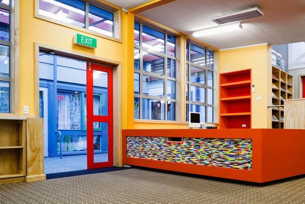 A brightly coloured commercial reception or entry area with a red and orange reception counter, a multicoloured mosaic-style front panel, open shelving, large framed windows, and a red glazed exit door. The space has patterned carpet, yellow painted walls, timber trims, and a suspended acoustic ceiling, creating a playful contemporary interior.