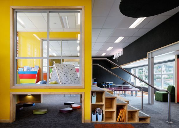 A brightly coloured contemporary educational or commercial interior, likely a school common area or activity space. The scene features vivid yellow walls and window framing, an internal glazed opening into a play or learning room, black walls, a suspended acoustic ceiling, linear recessed lighting, carpeted floors, tiered timber shelving integrated beneath a staircase, and scattered low circular seating. Large windows bring in daylight, while colourful furnishings and climbing or activity elements are visible through the internal glazing.
