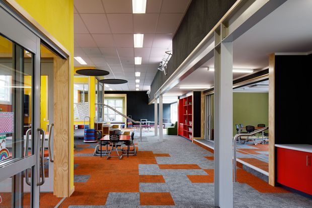 A bright, open-plan commercial interior with a playful, contemporary fit-out. The space features vivid yellow and red joinery, exposed structural columns, extensive internal glazing, open shelving, café-style tables and chairs, and a patterned carpet in orange and grey geometric blocks. A suspended linear lighting grid and visible upper-level or mezzanine-like circulation add to the office or educational workplace character.