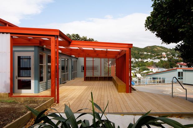 Exterior view of a contemporary residential outdoor entertaining area with a large timber deck, vivid red-orange steel or timber framing, translucent roof panels, grey horizontal cladding, and an integrated bench. A swimming pool and railing are visible to the right, with a hilly residential landscape beyond.