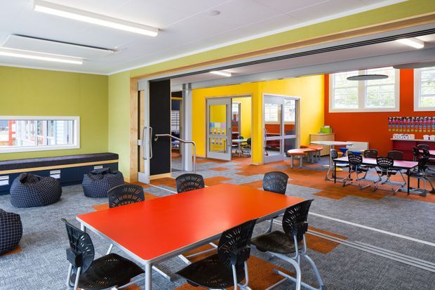 A bright, colourful communal interior resembling a school or childcare dining and activity area. The space features a large red rectangular table with black chairs in the foreground, additional tables and seating to the right, grey carpet with orange inset sections, vivid lime-green and yellow walls, and a red accent wall with shelving. Wide internal openings connect to smaller activity rooms, with a glazed entry or partition and exposed timber trim. Fluorescent linear lighting and acoustic ceiling panels create a functional institutional setting.