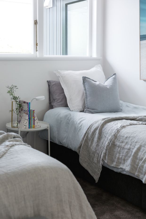 Bright, softly styled twin bedroom with two single beds dressed in pale blue-grey and white linens, layered pillows, and textured throws. A small round bedside table holds books, a slim lamp, and a delicate plant. White walls, white-painted window trim, and abundant natural light create a calm, airy atmosphere.