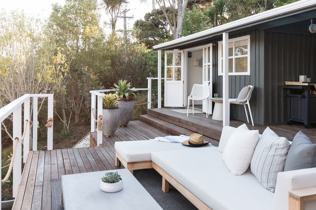 A relaxed outdoor deck and covered terrace beside a dark-clad compact dwelling, furnished with a large white outdoor sectional, low coffee table, two woven chairs, and a small bar or kitchenette. Timber decking, white railings, glazed doors and windows, and dense surrounding vegetation create a private, leafy setting.