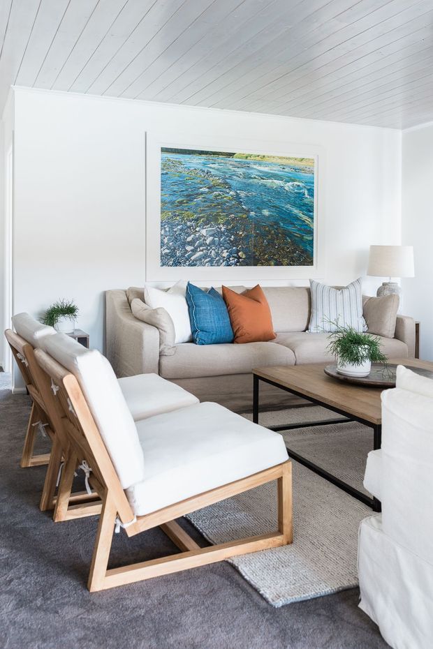 Bright, relaxed living room with white walls and a white-painted timber-lined ceiling. A neutral upholstered sofa and white-cushioned timber lounge chairs surround a low rectangular timber-and-black-metal coffee table on layered grey rugs. Blue and rust-orange cushions add colour, while a large framed image of a rocky blue-water landscape forms the main wall feature. Potted greenery and a floor lamp complete the room.