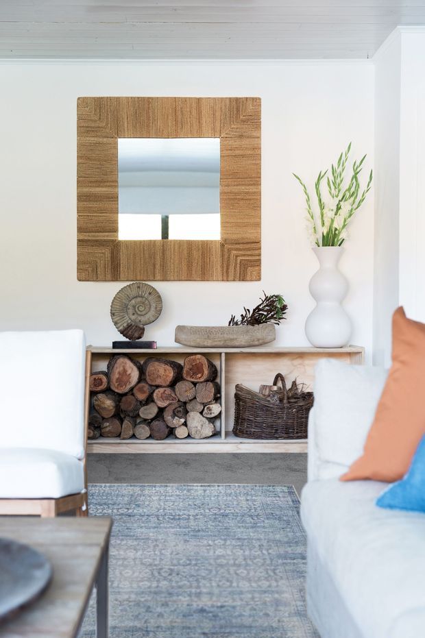 Bright, calm living room with white-painted walls and a white timber-lined ceiling. A pale timber console with open firewood storage and woven basket sits beneath a large square timber-framed mirror, styled with natural sculptural objects and a white vase of greenery. White sofas, a patterned blue-grey rug, and warm neutral accents create a relaxed coastal-natural feel.