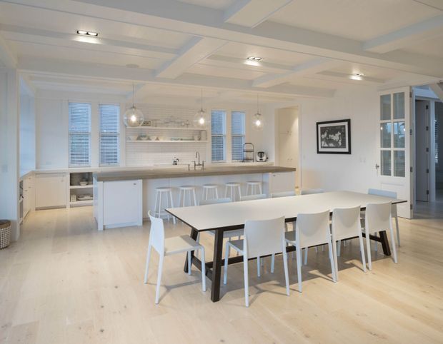 Bright, open-plan white kitchen and dining area with a long island featuring a timber benchtop and white cabinetry, pendant lights, integrated shelving, and a large rectangular dining table with white chairs. The room has pale timber flooring, white walls, extensive windows and glazed French doors, and a decorative coffered or beam-detailed ceiling. The overall design is clean, airy, and contemporary with restrained traditional detailing.