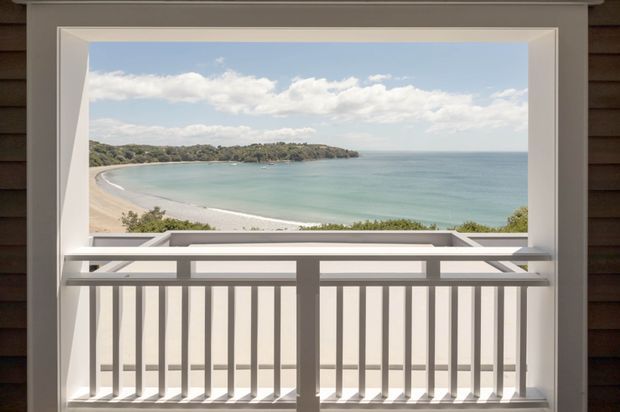 A framed ocean and beach view seen through a large opening, with a white-painted balustrade in the foreground and dark horizontal exterior wall cladding surrounding the opening. The scene is bright and coastal, with calm turquoise water, sandy shoreline, and a wooded headland beyond.
