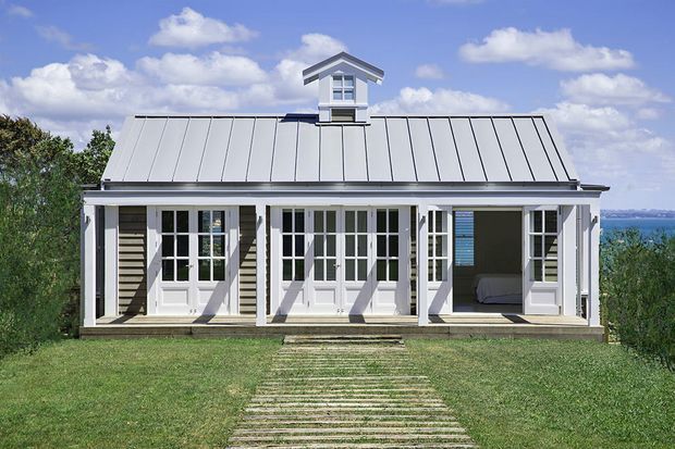 A compact, symmetrical coastal cottage viewed from the front, with a light weatherboard facade, broad covered veranda, repeated white French doors, and a standing-seam metal gable roof with a small central dormer. A grass lawn and simple stepping-stone path lead to the house, with water visible beyond the right side.