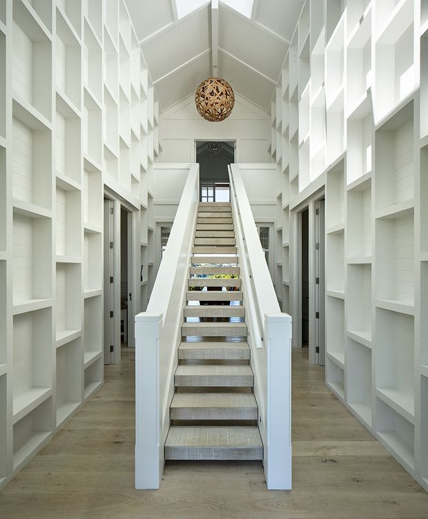 A bright, symmetrical interior hallway centred on a straight staircase leading to an upper landing. White built-in open shelving and geometric wall niches line both sides, with white painted timber wall panelling, a pitched vaulted ceiling, timber flooring, and a decorative spherical pendant light. Multiple doorways open off the corridor, creating a clean, airy architectural composition.