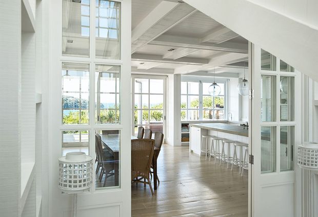 Bright, airy open-plan coastal-style interior viewed through white glazed partition doors. The space combines a dining area with wicker dining chairs and a large kitchen featuring a long island with white stools, extensive multi-pane windows, pale timber flooring, white wall and ceiling finishes, and pendant lighting. A blue water view and greenery are visible beyond the windows.
