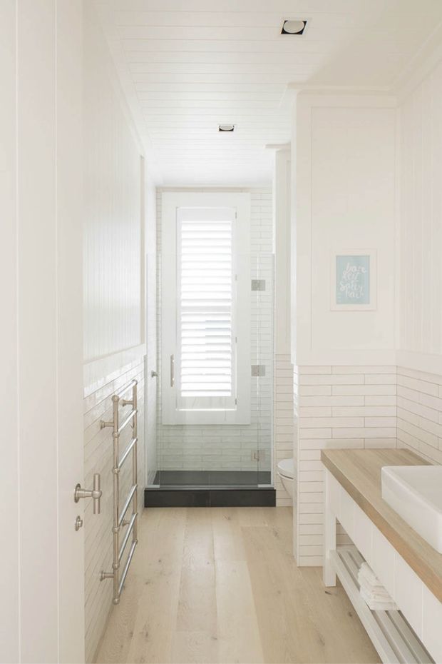Bright, narrow white bathroom with a glass shower enclosure at the end, a shuttered window, white horizontal wall tiling and panelling, pale timber-look flooring, and a vanity with a light timber benchtop and open towel storage. Brushed metal heated towel rails line the left wall, and recessed ceiling lights provide even illumination.
