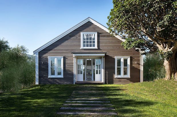 Front exterior view of a compact single-storey gable-roof house with dark horizontal weatherboard cladding, white trim, symmetrical multi-pane windows, and a glazed central entry porch. The house sits in a landscaped grassy setting with stepping-stone access and mature trees.