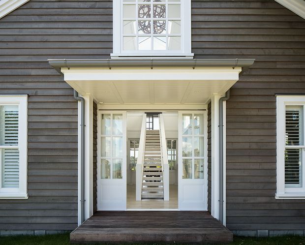 Symmetrical exterior view of a dark weatherboard house entry, featuring a covered porch with white trim, glazed double doors, sidelights, and a central staircase visible through the entrance. White-framed windows with shutters and a decorative upper window add a refined traditional character.