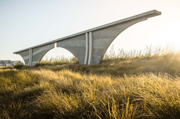 A sculptural elevated concrete structure with sweeping arched supports spans a grassy coastal-looking landscape. Warm low sunlight highlights the tall golden grasses, while the long minimalist form creates a strong horizontal silhouette against a pale sky.