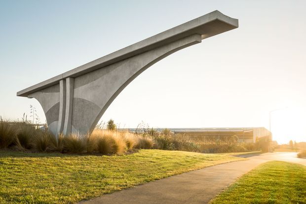 Exterior architectural landscape photograph featuring a sculptural curved concrete gateway or canopy structure beside a paved pedestrian path. The minimalist structure has a broad projecting horizontal roof slab supported by a sweeping arched concrete form, set within landscaped grass and ornamental grasses in warm low sunlight.