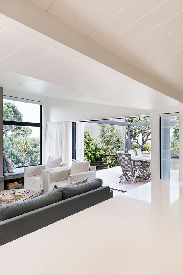 Bright open-plan living area with white painted walls and a white panelled ceiling, large black-framed windows, and wide sliding glass doors opening to a timber deck and outdoor dining area. A grey sofa, cream armchairs, and a low woven coffee table create a relaxed contemporary coastal feel, with abundant greenery visible outside.