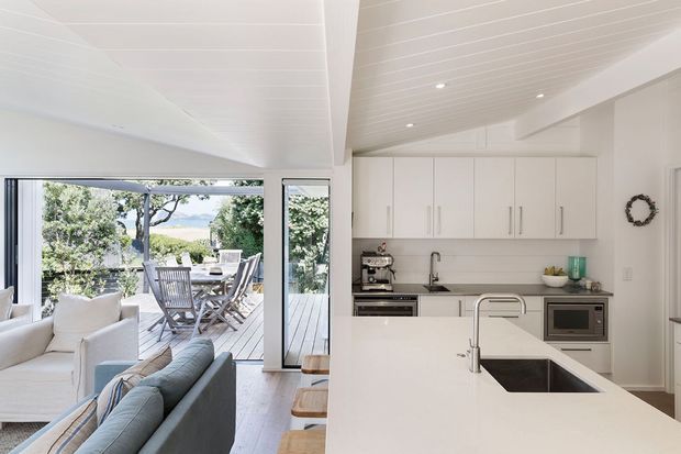Bright open-plan coastal-style interior showing a white kitchen with a large island and undermount sink, integrated appliances, white cabinetry, tiled splashback, and a dining deck visible through expansive glazing. A portion of the adjacent living area with sofas is visible in the foreground. The space has a white-lined ceiling, light timber flooring, and abundant natural light.