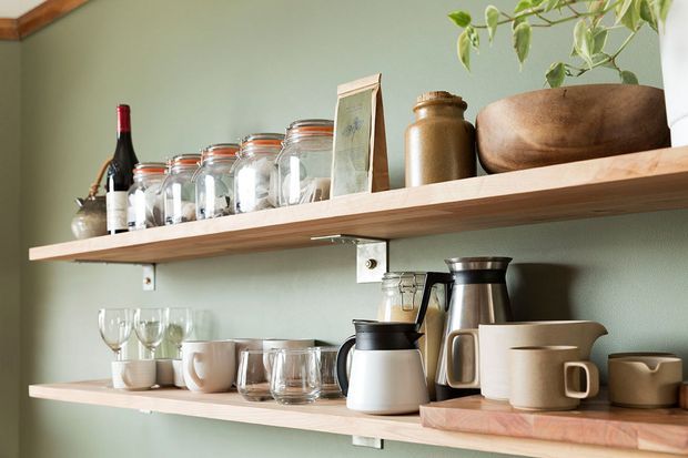 Close-up of two timber open shelves mounted on a muted sage-green wall, displaying glass jars, a wine bottle, ceramic mugs and cups, coffee and tea-making vessels, wooden containers, and a small trailing plant. The composition suggests a styled kitchen or pantry storage area with warm natural materials and a calm, earthy palette.