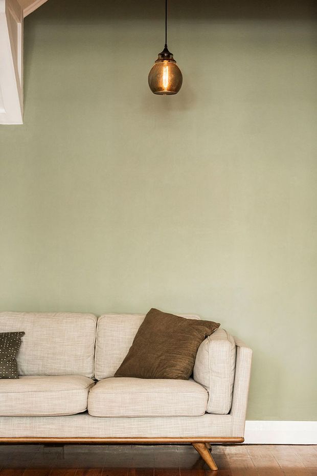 A close-up interior view of a mid-century-inspired upholstered sofa against a muted pale green wall. The room features a warm timber floor, white skirting, simple white ceiling trim, and a suspended amber glass pendant light. Neutral linen upholstery and earthy brown cushions create a calm, understated setting.