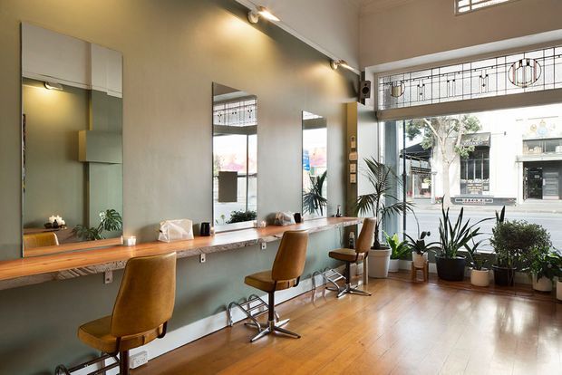 A bright, spacious hair salon interior with a long timber styling counter, wall-mounted mirrors, vintage brown salon chairs, polished timber flooring, indoor plants, and large street-facing windows with decorative leadlight transoms. Track lighting and exposed decorative wall elements contribute to a refined contemporary-retro commercial fit-out.
