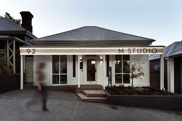 Street-facing single-storey studio or commercial frontage with a symmetrical white weatherboard facade, corrugated hipped roof, central glazed entrance, large windows, covered entry canopy, and prominent signage reading “92 M STUDIO.” A dark raised planter borders the entrance, with neighbouring buildings visible on either side.