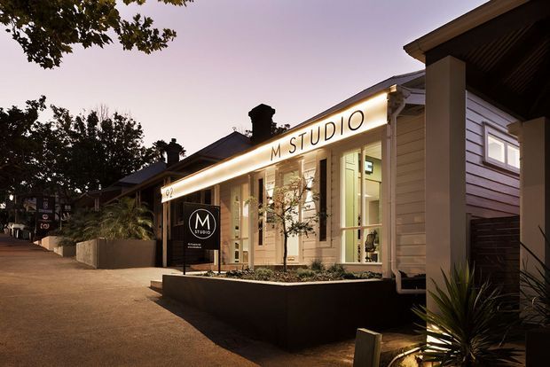 Dusk view of a small contemporary studio or commercial design showroom with illuminated M Studio signage, a light horizontal-clad facade, tall glazed entrance windows, dark roof elements, and landscaped raised planters along a paved approach.