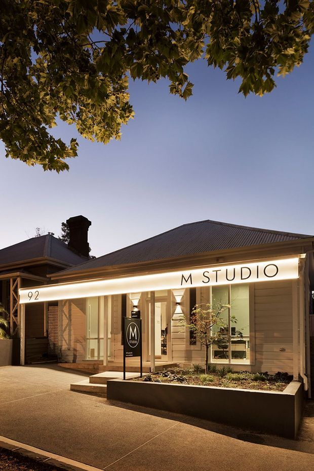 Exterior dusk photograph of a low-rise commercial studio occupying a traditional residential-style building. The facade has pale horizontal cladding, large illuminated windows, a prominent projecting sign reading “M STUDIO,” a dark corrugated pitched roof, and a small landscaped raised planter along the pavement. Mature tree branches frame the upper edge of the image.