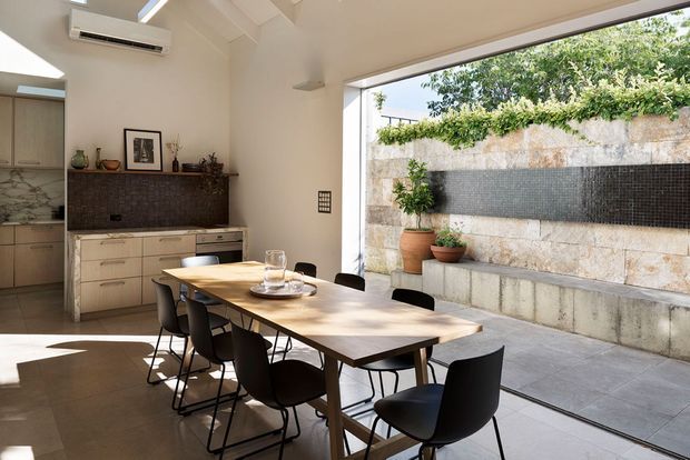 Bright contemporary open-plan dining and kitchen space with a large timber dining table and black dining chairs beside full-height glazing. The room opens onto a paved courtyard with a stone and dark tiled feature wall, built-in bench, and potted plants. Neutral cabinetry, pale walls, and exposed ceiling beams create a clean, refined interior.