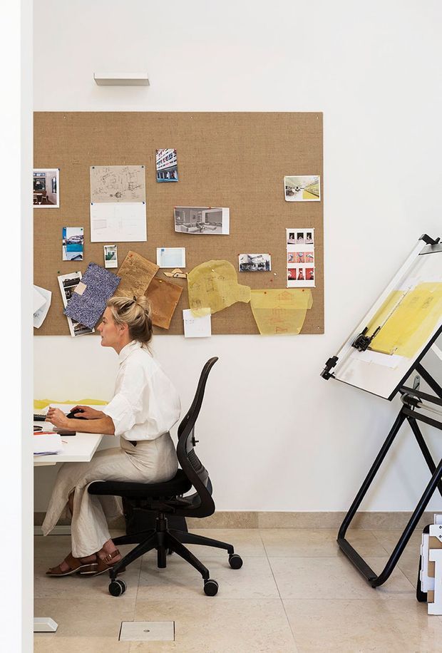 A bright, minimalist home office or design studio with a white desk, an occupied ergonomic office chair, a large cork inspiration board covered with plans, images and material samples, and a drafting table on an easel. The room has pale tiled flooring, white walls, and a clean, functional creative-workspace atmosphere.