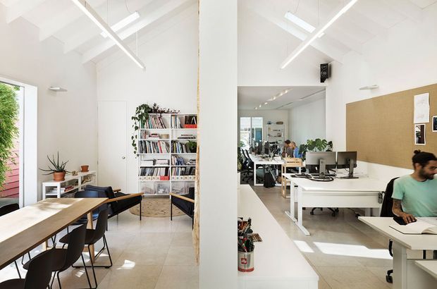 Bright open-plan creative studio combining a dining or meeting area with a shared office workspace. The predominantly white interior has exposed high ceilings, linear pendant lighting, tiled flooring, timber tables, open shelving filled with books, a large corkboard, desks with computers, and abundant natural light.