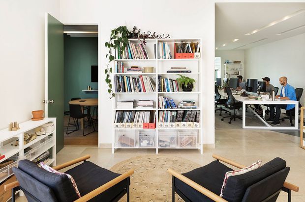 Bright, open-plan office lounge with two black leather-and-wood lounge chairs in the foreground, a large white shelving unit filled with books and plants, and a shared workspace visible through the opening on the right. A small adjoining meeting or dining area is visible through the doorway on the left. The space has pale walls, light flooring, and abundant natural light.