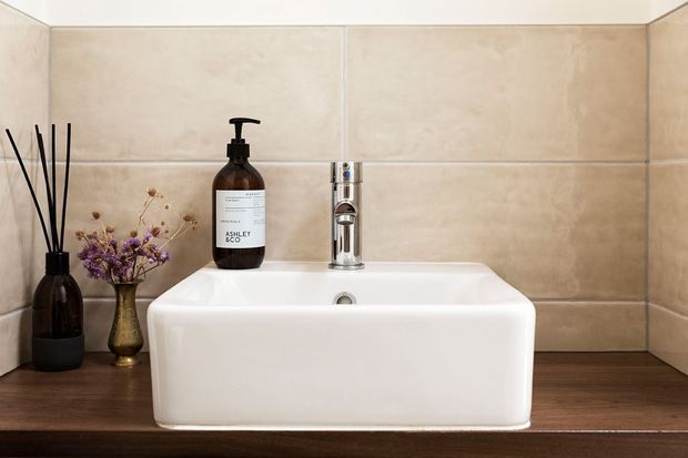 Close-up of a compact bathroom vanity featuring a white vessel basin with chrome mixer tap, set against beige tiled walls and a dark timber-look countertop. Decorative reed diffuser, soap dispenser, and small dried floral arrangement add restrained styling.