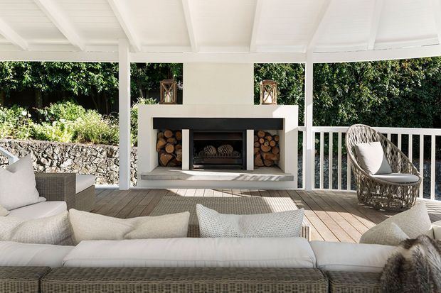 Bright covered outdoor lounge with a white-painted timber ceiling and posts, timber decking, a large built-in masonry fireplace, woven outdoor seating, and lush garden screening beyond the balustrade. The space has a relaxed coastal-resort feel with neutral furnishings and natural stone and wood details.