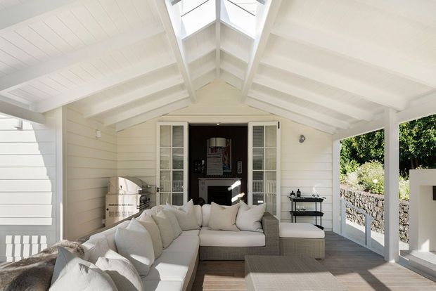 Bright, airy covered outdoor lounge or sunroom with white-painted weatherboard walls, exposed pitched roof framing, and a central skylight. The space is furnished with a large light-coloured sectional sofa and ottoman, with glass doors opening toward a landscaped exterior and a darker interior room beyond.