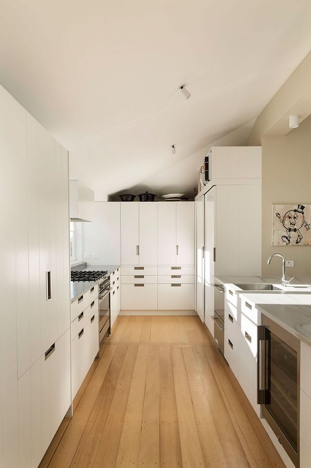 A narrow, elongated galley kitchen with extensive white built-in cabinetry, integrated appliances, a marble-look island or peninsula with sink, stainless-steel tapware, and warm timber floorboards. The sloping ceiling and bright natural light create an airy, streamlined space.