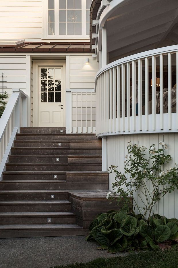 Exterior view of a refined residential entry with broad dark timber steps, white painted weatherboard cladding, a glazed front door, and a curved verandah or balcony with closely spaced white balustrades. The composition includes layered roof details, traditional window proportions, and planted garden beds beside the stairs.