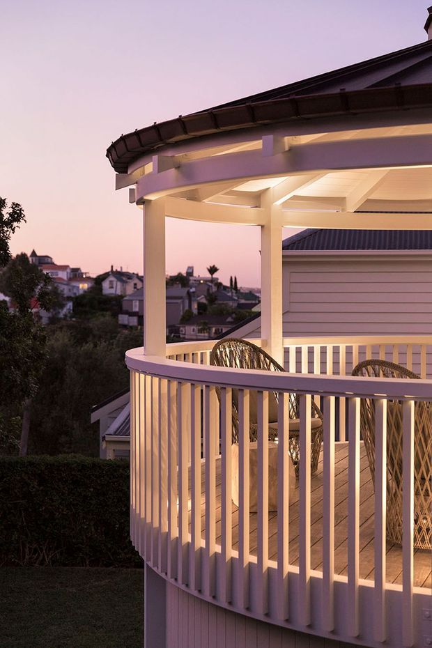 A dusk photograph of an elevated residential outdoor deck or balcony with a curved white-painted balustrade, covered pergola, exposed roof framing, warm integrated lighting, and wicker seating. A suburban neighbourhood is visible in the background.