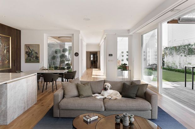 Bright open-plan contemporary living space with a large grey sectional sofa in the foreground, opening onto a dining area and kitchen. Full-height glazing connects the room to a timber deck and landscaped courtyard. The interior features pale timber flooring, white walls and joinery, a stone kitchen island, and curated artwork and furnishings.