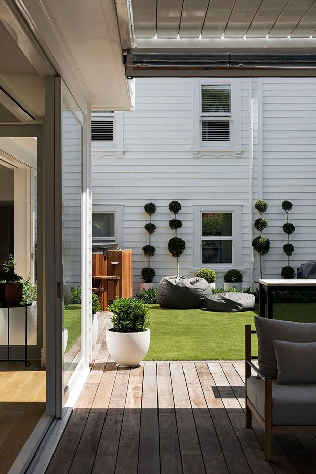 A bright, refined courtyard viewed from a covered timber deck through open glazed doors. The courtyard is enclosed by white horizontal weatherboard walls with decorative window trims, topiary plants, artificial or closely clipped lawn, and beanbag seating. A timber outdoor bar or cabinet and dark-framed outdoor furniture complete the space. The image also shows a glimpse of the adjacent interior and a partially screened overhead structure.