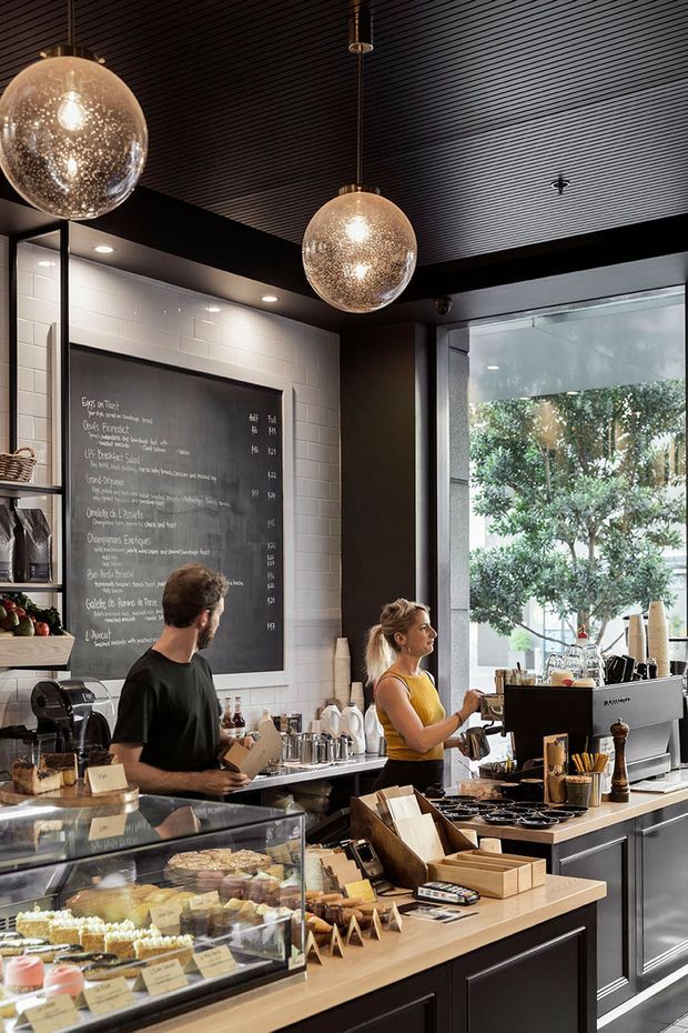 A stylish café interior with a service counter, pastry display case, commercial espresso machine, chalkboard menu, open shelving, and staff working behind the counter. The space features dark ribbed ceiling panels, black structural beams and cabinetry, white subway-tile walls, warm timber counters, and large glazed frontage with greenery visible outside. Spherical pendant lights add a decorative focal point above the bar.