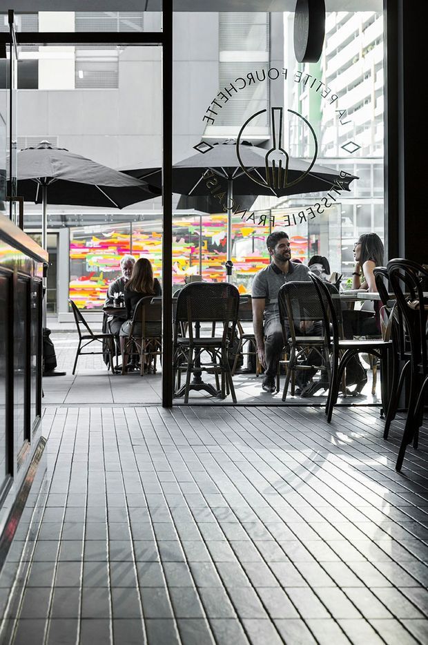 A bright urban café or restaurant interior viewed through large black-framed glazed doors. The space has dark rectangular tiled flooring, black café tables and chairs, outdoor umbrellas, and several diners. A colorful abstract mural is visible across the courtyard, with surrounding multi-storey buildings creating a dense city setting. Café lettering and a circular logo appear on the glass.