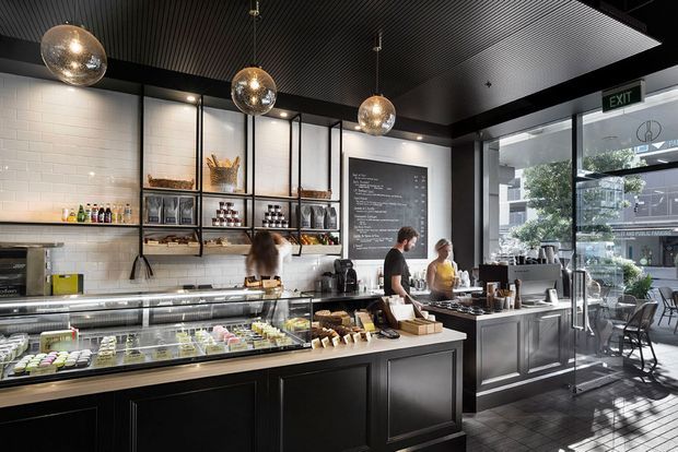 A refined contemporary café interior with a long dark service counter and refrigerated pastry display, open shelving stocked with café supplies, white subway-tile walls, a ribbed dark ceiling, and three spherical pendant lights. Staff work behind the counter, while full-height glazing opens the café visually to the street outside.